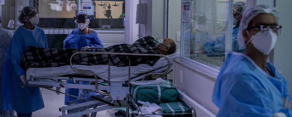 Em corredor hospitlar, mulher paramentada com vestes de proteção e máscara observa a câmera diante de janela de vidro, com homem coberto comum cobertor xardrez em maca e dois enfermeiros atrás