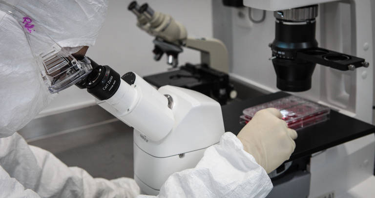 Pessoa com roupa protetora branca observa lâmina em microscópio