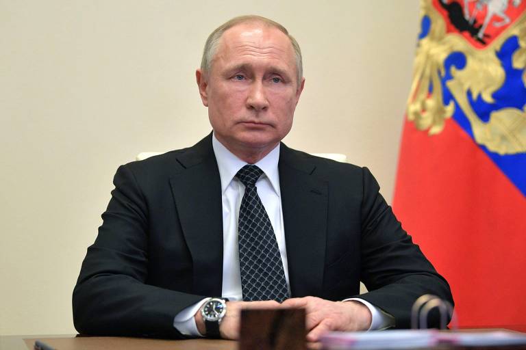 Putin está sério, sentado em frente a uma mesa, com paletó preto e camisa branca. Atrás está a bandeira da Rússia com o brasão de armas
