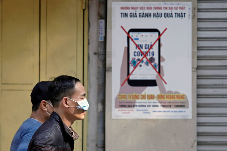 Moradores com máscaras de proteção passam em frente a cartaz que alerta contra a divulgação de notícias falsas sobre o coronavírus em Hanói, no Vietnã
