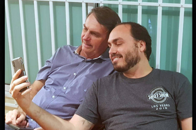 De camisa de botões azuis, Jair Bolsonaro, branco e de cabelos curtos grisalhos, vê algo no telefone de Carlos, branco, de barba e cabelos curtos escuros e camiseta cinza