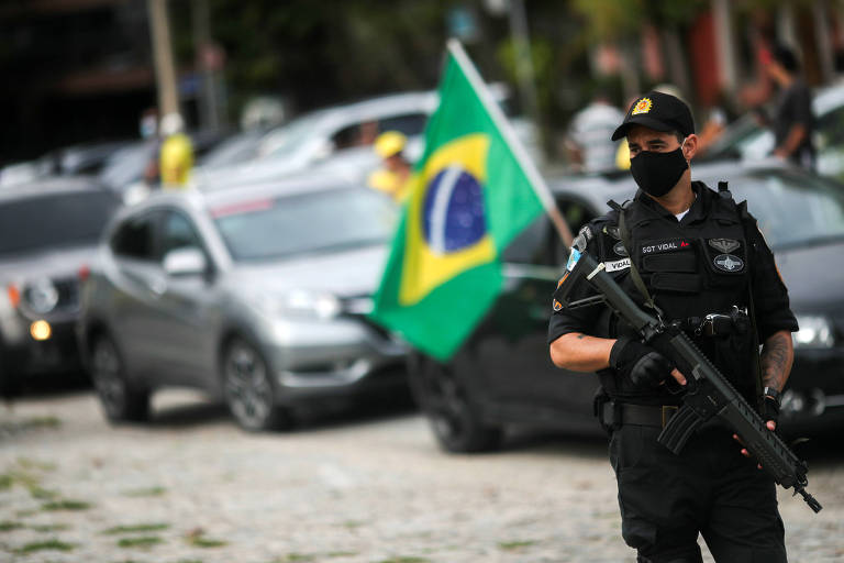Policial carregando fuzil e usando máscara e carros com bandeira do Brasil ao fundo