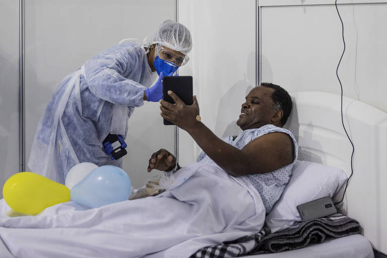 Homem deitado em cama hospitalar mostra tablet para enfermeira paramentada, com bexigas amarelas e azuis sobre a cama