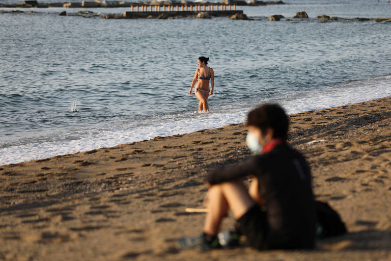 uma pessoas está sentada na praia, de máscara e roupa preta. ao fundo, uma mulher banha-se no mar
