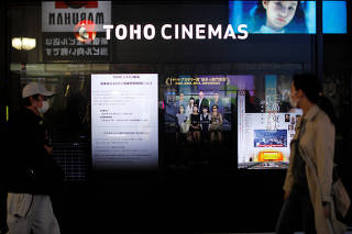 Passersby wearing protective face masks walk past notices of closed and business hours reduction displayed in front of a movie theater in Tokyo