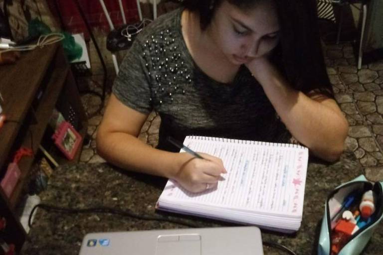 Menina sentada à mesa escreve no caderno escolas com notebook na frente dela
