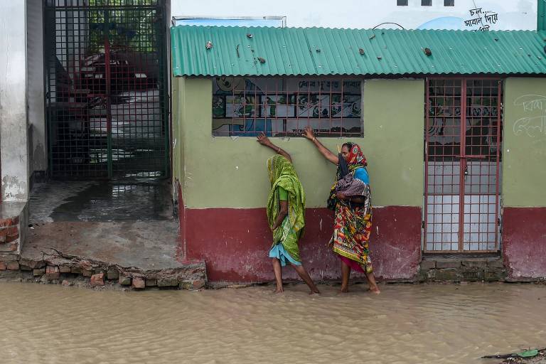 Duas mulheres agarram o peitoril da janela de um estabelecimento. Seus pés estão submersos na água que alaga a rua 