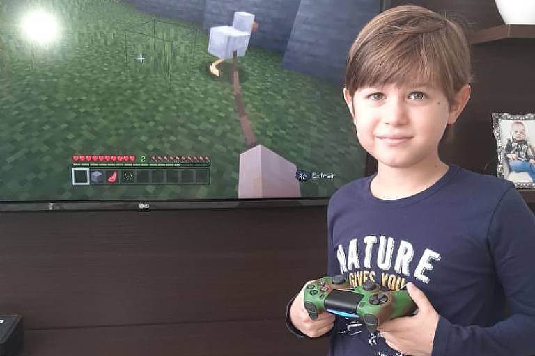 Menino veste pijama e está em pé, na frente da televisão, segurando o controle de um console 