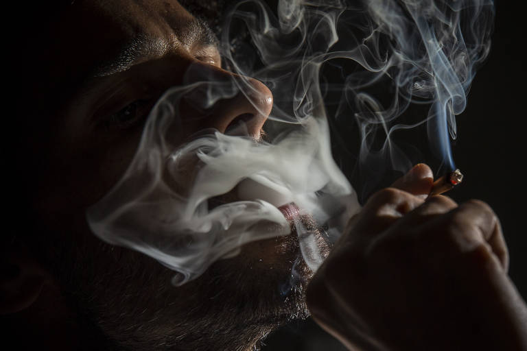 Fotografia colorida foca o rosto de um homem branco fumando maconha; o ambiente está escuro e não dá para saber a identidade dele; a fumaça do cigarro cobre parte do rosto
