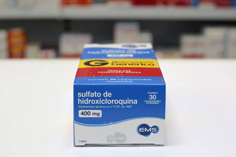 Caixa do medicamento hidroxicloroquina vista de baixo - a embalagem está em cima de uma mesa branca com prateleira de medicamentos ao fundo desfocada