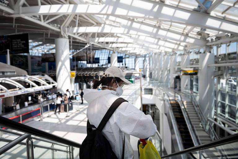 Mulher com roupa branca, máscara e boné desce escada rolante em terminal de aeroporto 