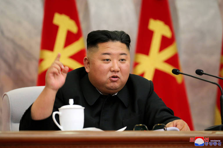 Kim Jong Un está sentado em frente a uma mesa, há bandeiras do partido atrás dele, ele está de roupa preta, levanta o dedo indicador enquanto fala e há um pequeno bule branco na mesa