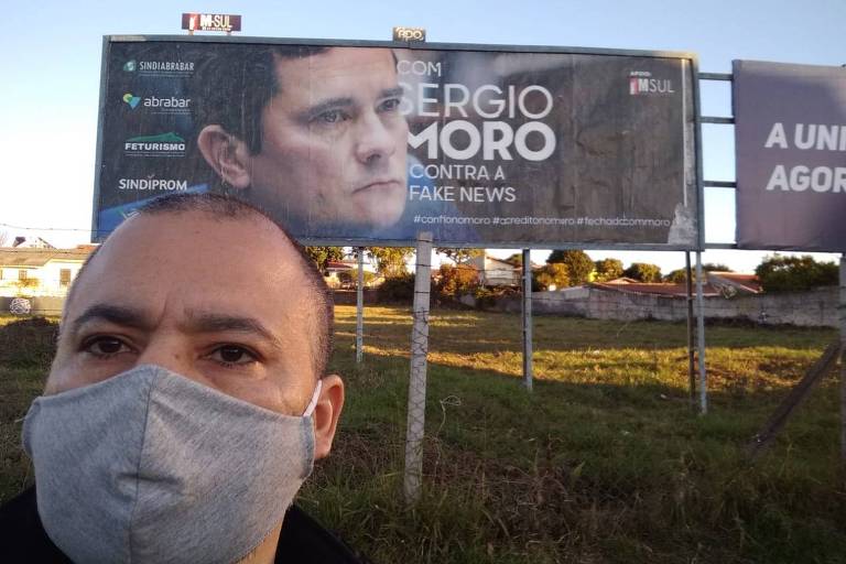 Homem de máscara em frente a outdoor com foto de Sergio Moro