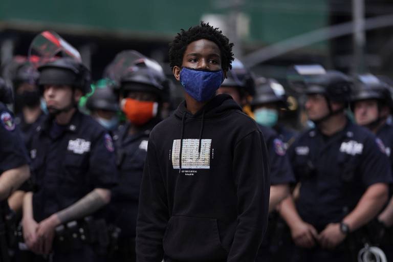 Um menino negro, de máscara azul e moletom preto está parado em frente a uma filha de policiais com uniformes pretos. Alguns deles usam máscaras