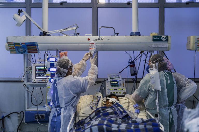 Imagem mostra ambiente de UTI, com leito, equipamentos diversos e dois profissionais de saúde cuidando de um paciente