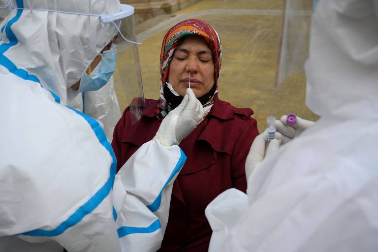 enfermeiros paramentados em roupas de proteção brancas colocam cotonete no nariz de uma idosa, que é branca e usa lenço colorido na cabeça