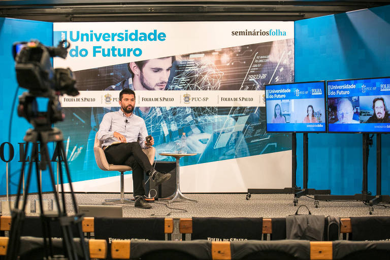 Fabio Takahashi aparece sentado em um palco, na frente do banner do seminário; ao seu lado, dois monitores mostram os convidados que acompanham o evento online