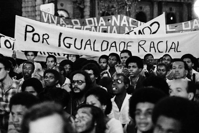 Foto em preto e branco, com dezenas de pessoas, a maioria delas negras, parte delas segura cartazes e em destaque aparece o que está escrito "por igualdade racial"