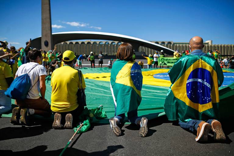 Manifestantes a favor do presidente Bolsonaro se ajoelham durante protesto com uma bandeira brasileira, próximo ao quartel-general do exército, contra a decisão do governador do Distrito Federal, Ibaneis Rocha (MDB), de impedir que multidões participem de manifestações em meio à pandemia de coronavírus, em Brasília.
