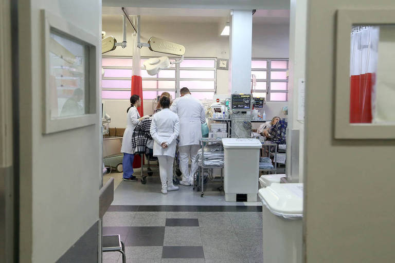 No Paraná, taxa de ocupação de leitos para Covid-19 cresceu em poucos dias; na foto vemos várias pessoas de costas, em torno de um leito, visto a partir de um corredor do Hospital do Trabalhador, em Curitiba
