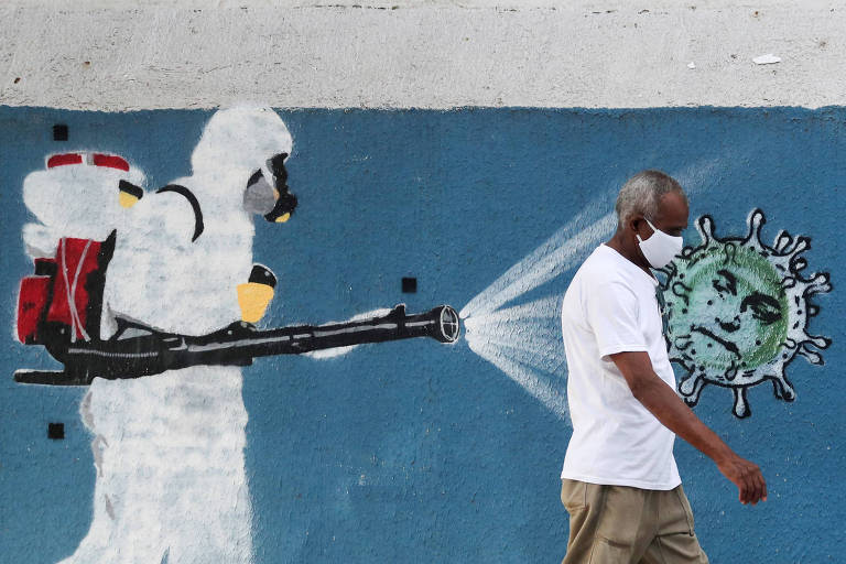 Homem caminha em frente a grafite em que homem com equipamento de proteção desinfeta vírus com rosto do presidente Jair Bolsonaro
