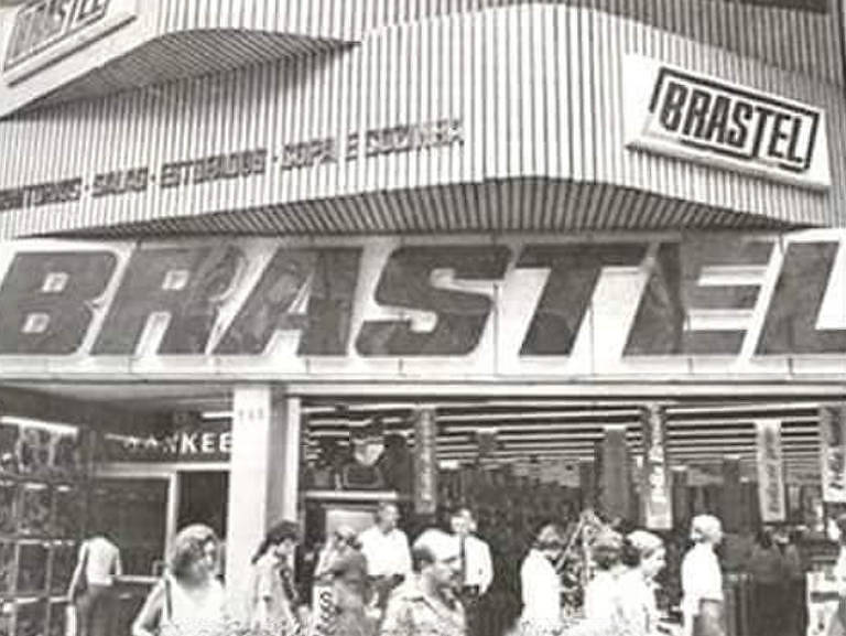 A imagem em preto e branco mostra a fachada da loja Brastel na década de 1970. Na frente da loja, há várias pessoas