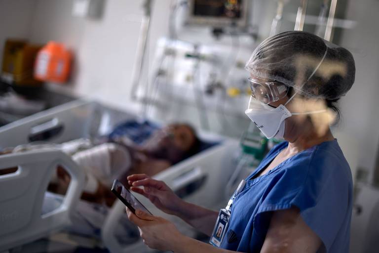 Imagem mostra profissional da saúde vestida de camisa azul, com máscara e touca. Ela segura uma prancheta. Ao fundo, há um paciente deitado em uma maca, em um quarto de hospital.