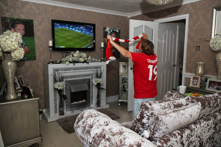 A imagem mostra uma pessoa em uma sala de estar assistindo a um jogo de futebol na televisão. A pessoa está vestindo uma camiseta vermelha e segurando um cachecol do Liverpool. O ambiente é decorado com paredes marrons, um sofá claro e uma lareira decorativa. Na parede, há uma televisão exibindo uma partida de futebol.