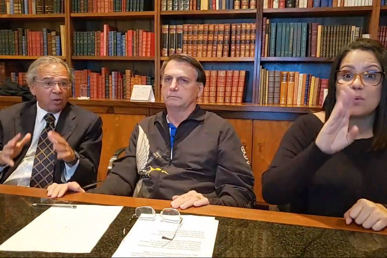 Paulo Guedes e intérprete de libras gesticulam, enquanto Bolsonaro olha para frente 