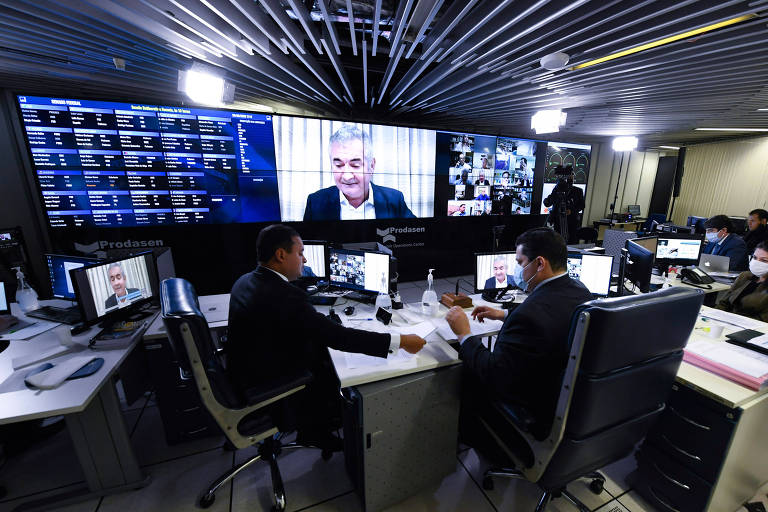 Dois homens sentados com monitor de videoconferência ao fundo