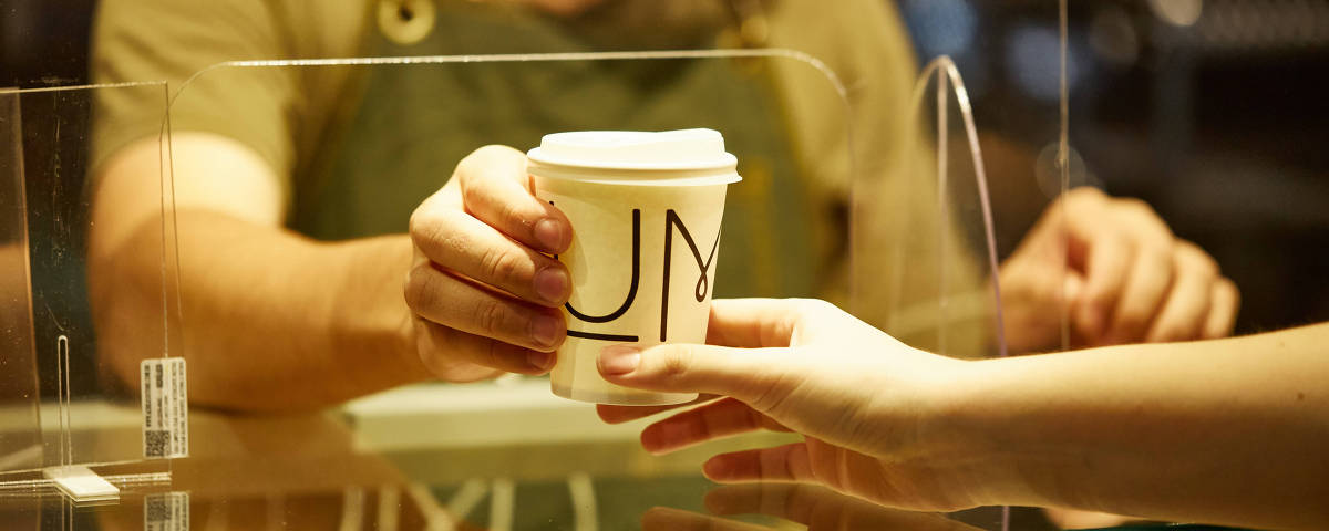 Cliente recebe café através de placa transparente que evita o contato com o barista na cafeteria Um Coffee Co.