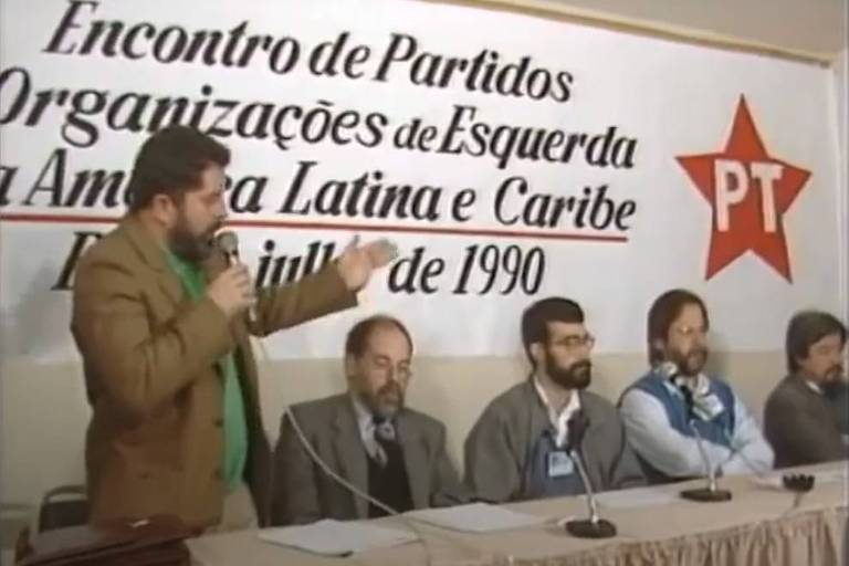 Lula, de cabelo e barba castanhos, está em pé, à esquerda da foto, segurando microfone e atrás de mesa. Ao lado dele, em direção à direita da foto, estão outros homens, sentados. Atrás deles, um painel onde se lê em itálico "Encontro de Partidos Organizações de Esqueda x América Latina e Caribe - julho de 1990" e a estrela do PT, vermelha com a sigla PT em branco no centro