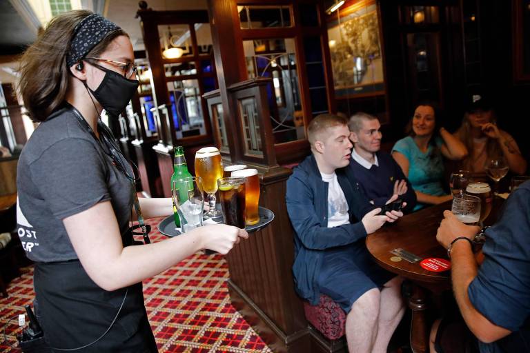 Funcionária de máscara serve clientes aglomerados em mesa de pub londrino