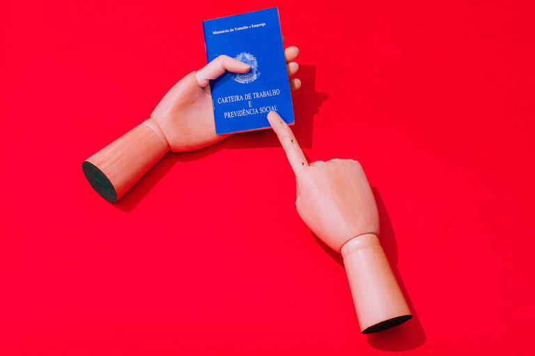 Mão esquerda de madeira seguram a carteira de trabalho, com capa azul; o fundo é vermelho. A mão direita aponta com o dedo indicador o que está escrito, que é CTPS