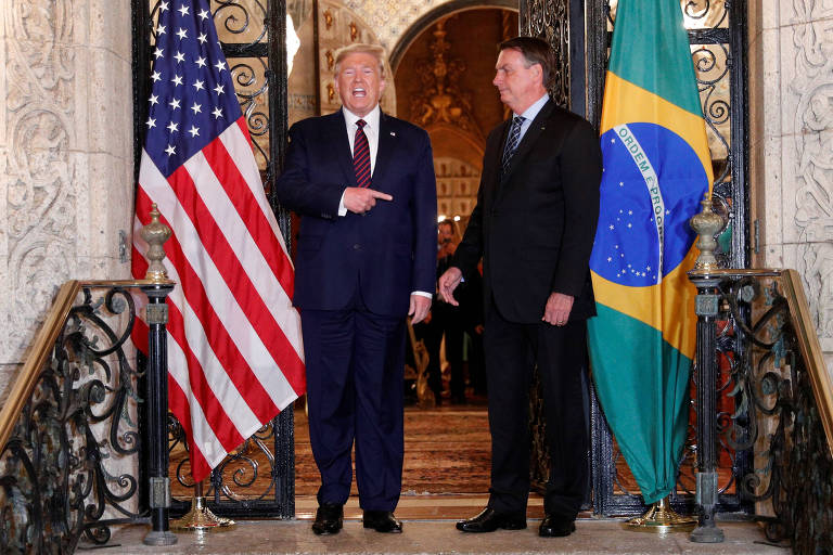 De pé e sorrindo, o então presidente dos Estados Unidos, Donald Trump, aponta para Jair Bolsonaro, que está a seu lado, durante encontro em Palm Beach, na Flórida; ao lado de Trump há uma bandeira dos Estados Unidos e ao lado de Bolsonaro há uma bandeira do Brasil