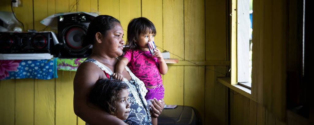 Mulher com menina pequena ao colo e outra um pouco maior ao lado, em uma sala de ripas de madeira amarela, observa movimento da janela, iluminada pelo sol