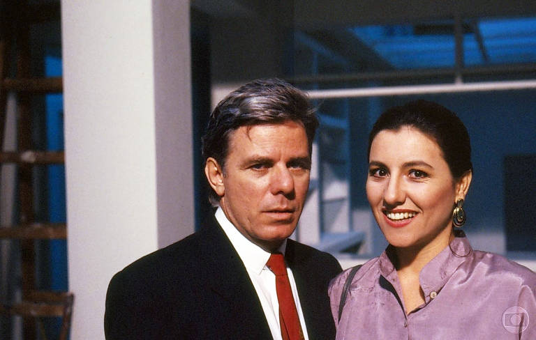 A imagem mostra um homem e uma mulher posando juntos. O homem tem cabelo grisalho e usa um terno escuro com uma camisa branca e gravata vermelha. A mulher tem cabelo escuro e usa uma blusa rosa. Ambos estão em um ambiente com paredes brancas e uma iluminação azul ao fundo, que sugere um espaço moderno.