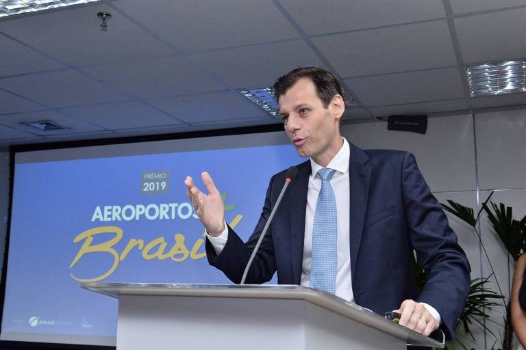 Chefe da secretaria da aviação civil, Ronei Glanzmann, fala em um púlpito. Atrás, uma apresentação em que se lê o slide "Aeroportos Brasil" em fundo azul