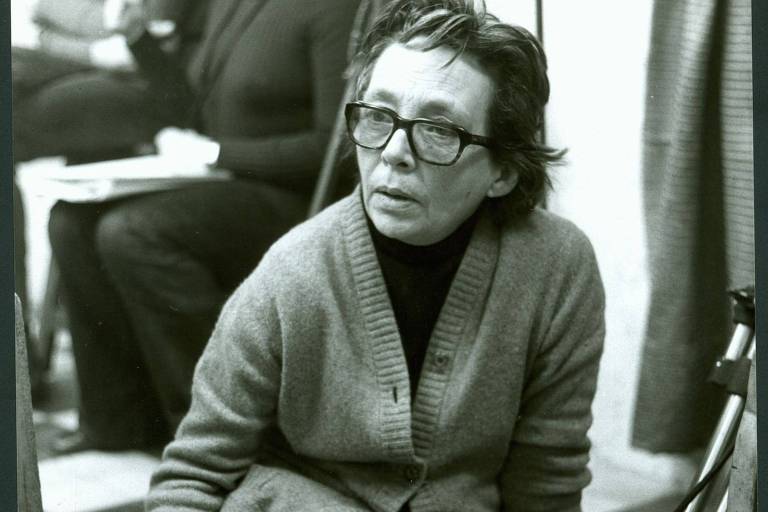 Marguerite Duras veste saia e casaco de lã. De óculos, ela está sentada no chão com as pernas cruzadas