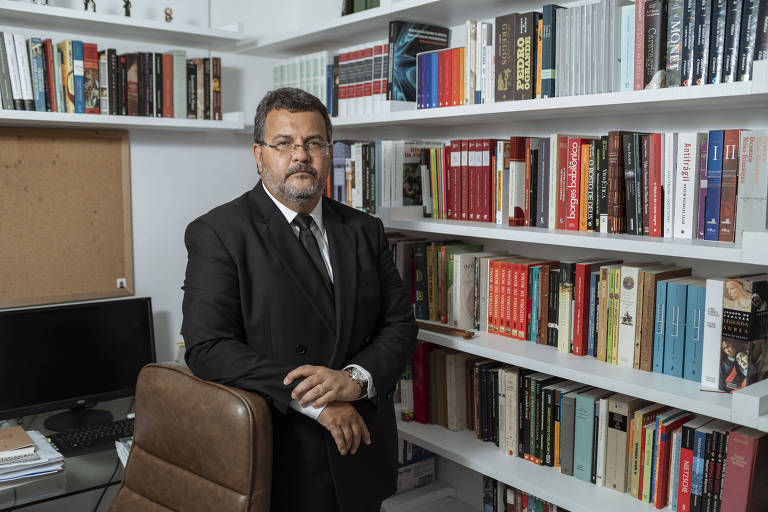 Rodrigo, um homem branco, de barba grisalha, veste terno escuro e está em pé no canto de um escritório, entre uma estande de livros, ao fundo, e ao lado da cadeira e computador 