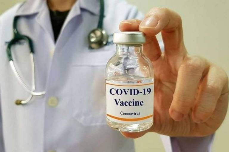Pessoa com avental branco e estetoscópio no pescoço segura frasco com etiqueta com escrito "Covid-19 Vaccine"