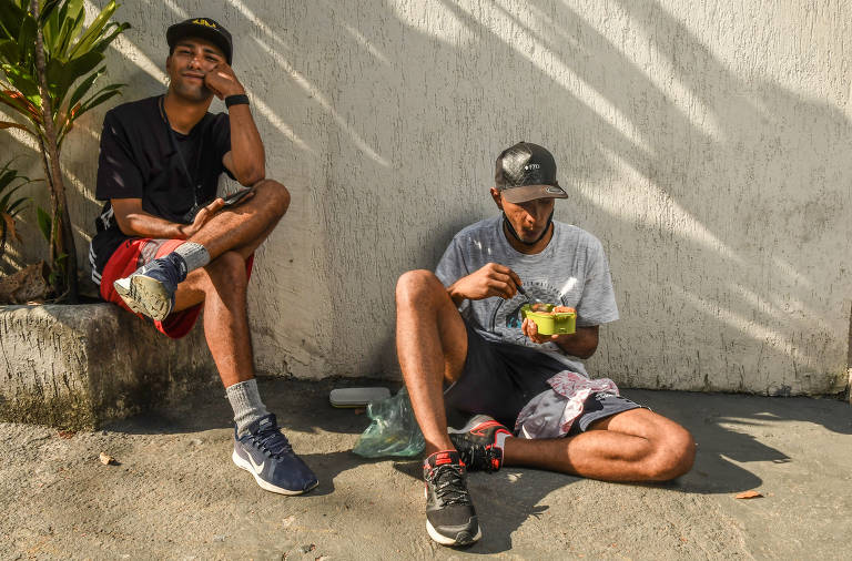 Guilherme Carvalho dos Santos, 30, come sentado na calçada ao lado de uma lanchonete em uma das esquinas da avenida Rebouças; arroz, feijão e frango; tudo frio. Ele pedala da Raposo Tavares até ali todos os dias. Ao seu lado, Gabriel Santana, 25, espera a fome bater para comer sua marmita também fria