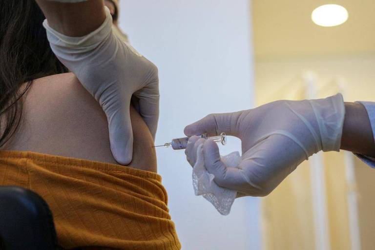 Detalhe do ombro de mulher recebendo vacina por injeção dada por mãos com luvas brancas