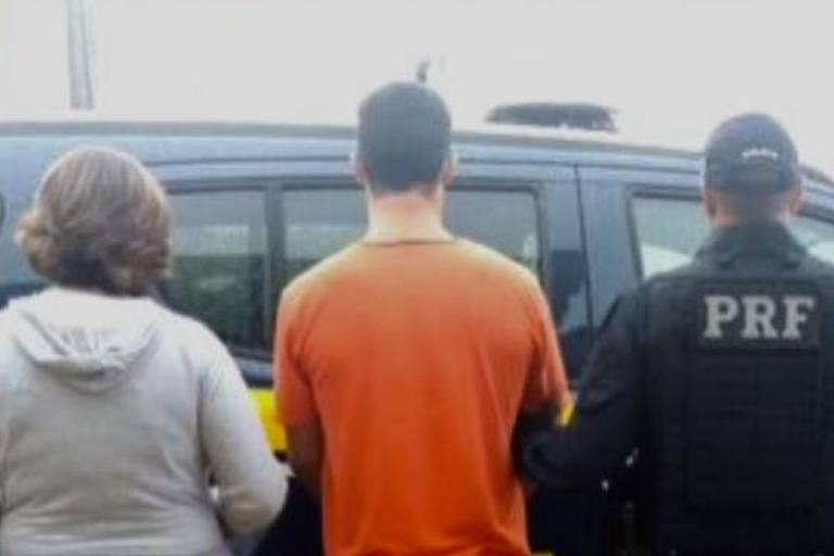 De costas estão um homem de camisa laranja entre dois policiais, um deles usando um colete com a sigla da Polícia Rodoviária Federal.