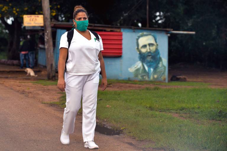 Mulher com uniforme branco e máscara caminha por rua com painel com Fidel Castro ao fundo