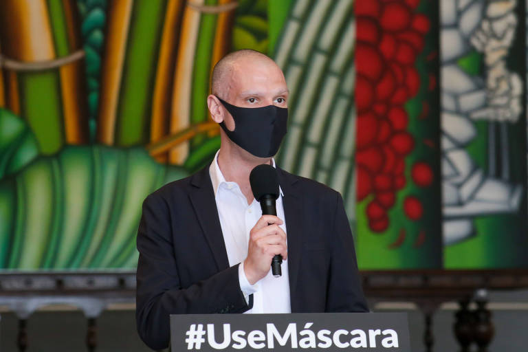 Bruno Covas, um homem branco e careca, está de pé em frente a um púlpito com a inscrição "#UseMáscara". Ele está de terno preto, camisa branca e máscara. Ele segura um microfone.