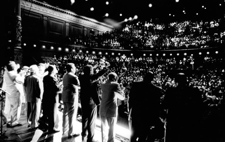 Um grupo de homens, enfileirado, está em cima de um palco cumprimentando a plateia de um teatro