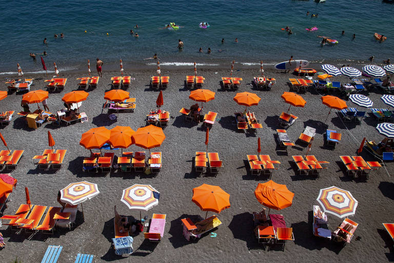 A imagem mostra uma praia com várias cadeiras e guarda-sóis laranja dispostos em um padrão organizado. Há pessoas na água e na areia, aproveitando o dia ensolarado. O mar é azul e calmo, e há também algumas toalhas e guarda-sóis brancos e listrados ao fundo.