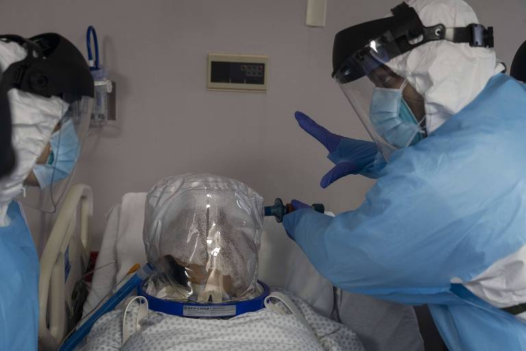 Ao centro, paciente acamado e com face shield sendo atendido por duas pessoas da equipe médica (uma à direita e, outra, à esquerda da cama) paramentadas contra o novo coronavírus