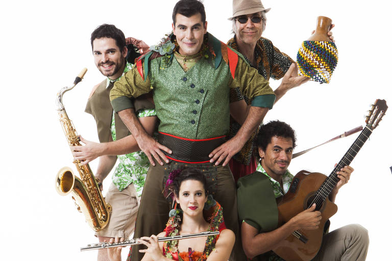 Grupo de quatro homens e uma mulher, fazendo pose segurando instrumentos musicais como flauta transversal, violão, sax e chocalho. Eles vestem roupas das cores verde, laranja, azul e vermelho, de estilo circense 